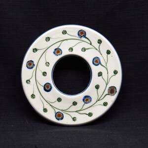 Könnte beinhalten: Ein weißer Keramik-Topfuntersetzer mit einem Ring in der Mitte. Der Ring ist von einem grünen Ranken mit blauen und braunen Blüten umgeben.