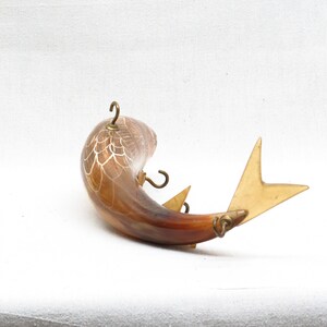 K&ouml;nnte beinhalten: Ein brauner und wei&szlig;er fischf&ouml;rmiger Wandhaken aus Horn mit goldenen Metallakzenten. Der Haken hat drei Haken an der Seite und eine Schwanzflosse.