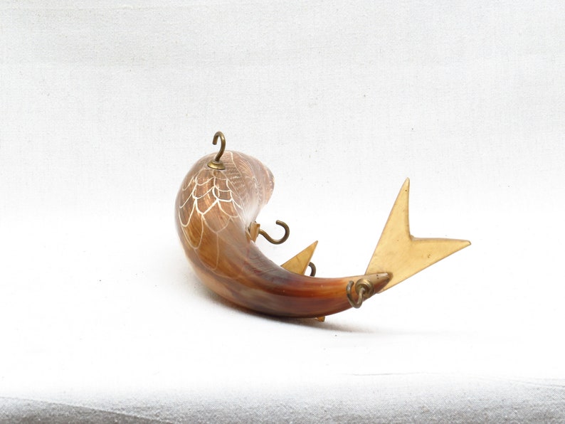 K&ouml;nnte beinhalten: Ein brauner und wei&szlig;er fischf&ouml;rmiger Wandhaken aus Horn mit goldenen Metallakzenten. Der Haken hat drei Haken an der Seite und eine Schwanzflosse.