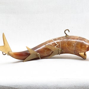 K&ouml;nnte beinhalten: Ein geschnitztes Horn in Form eines Fisches mit einem Messinghaken oben und Messingakzenten am Schwanz. Das Horn ist braun und wei&szlig; und hat ein detailliertes Design.