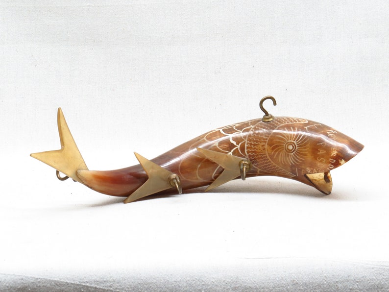 K&ouml;nnte beinhalten: Ein geschnitztes Horn in Form eines Fisches mit einem Messinghaken oben und Messingakzenten am Schwanz. Das Horn ist braun und wei&szlig; und hat ein detailliertes Design.