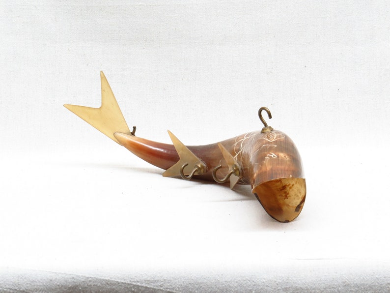 K&ouml;nnte beinhalten: Ein dekorativer, an der Wand montierter Horn mit einem Fischdesign. Das Horn besteht aus braunem und wei&szlig;em Material und hat drei Messinghaken. Der Fischschwanz besteht aus hellbraunem Material und hat eine wei&szlig;e Spitze.