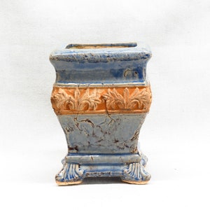 May include: A blue and orange ceramic planter with a square top and a decorative design around the middle. The planter has four feet.