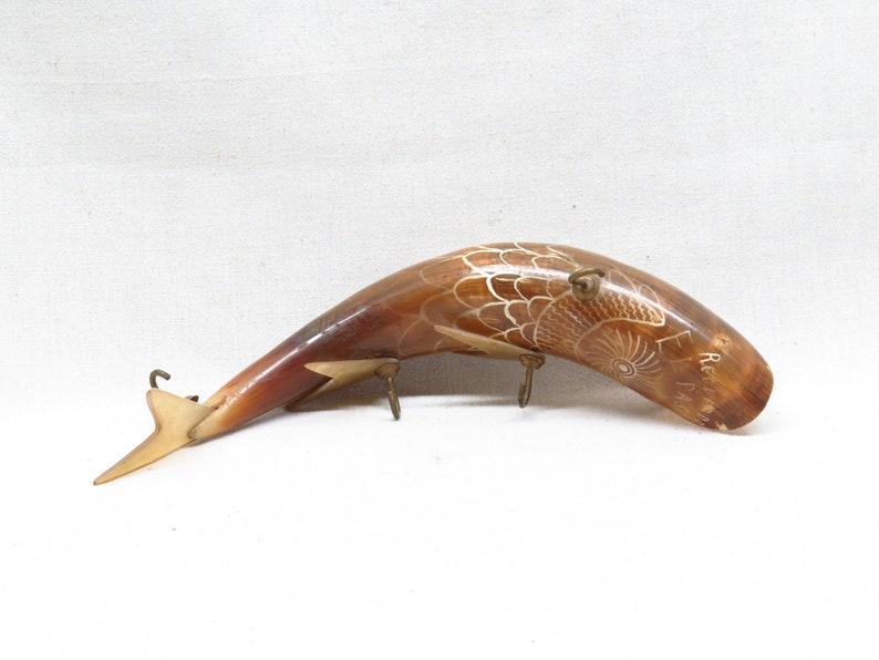 K&ouml;nnte beinhalten: Eine geschnitzte Hornskulptur eines Fisches mit einem detaillierten Design. Das Horn ist braun und das Design ist wei&szlig;. Die Skulptur ist auf einem Metallst&auml;nder mit drei Haken montiert.