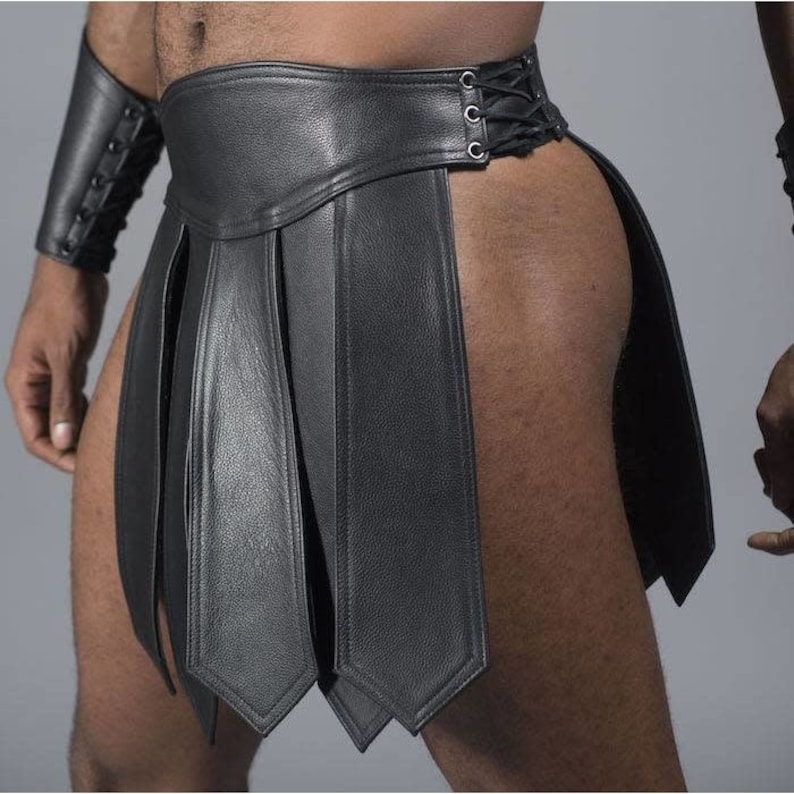 May include: Black leather gladiator skirt with vertical panels and a laced closure. The skirt features a wide waistband and is designed with a split on the side. A matching black leather arm cuff is also visible.