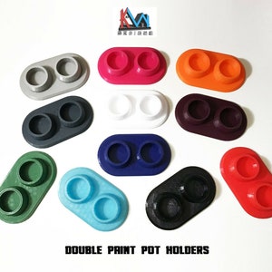 May include: Assortment of ten double paint pot holders in various colours, including grey, pink, orange, white, purple, blue, green, and black. The holders are oval-shaped with two circular indentations for paint pots. The text "DOUBLE PAINT POT HOLDERS" is printed below the holders.