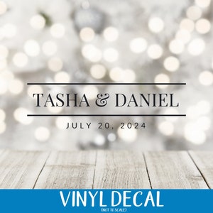 May include: A white and silver bokeh background with a black border and the text "TASHA & DANIEL" and "JULY 20, 2024". Below the text is a white wooden surface and a blue banner with the text "VINYL DECAL (NOT TO SCALE)".