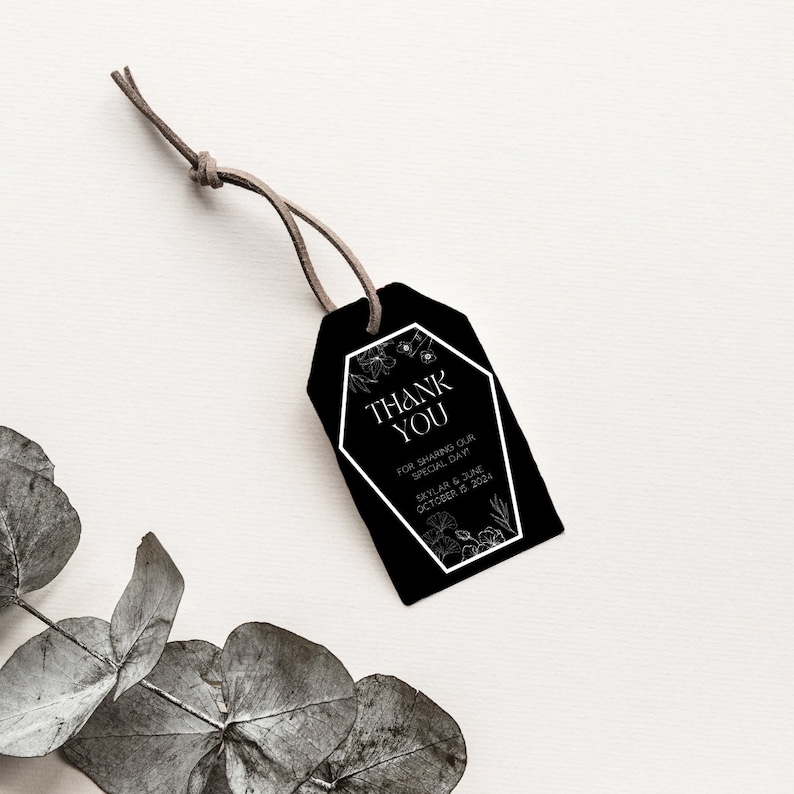 May include: Black and white wedding favour tag with a coffin-shaped design and floral accents. The text reads "Thank You" and "For sharing our special day."  The tag also includes the names "Skylar & June" and the date "October 22, 2024".
