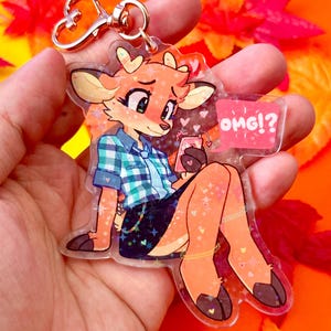 May include: A clear acrylic keychain featuring a cartoon deer character with a plaid shirt and skirt, holding a phone. The keychain has a gold-colored clasp and a speech bubble that says "OMG!?"