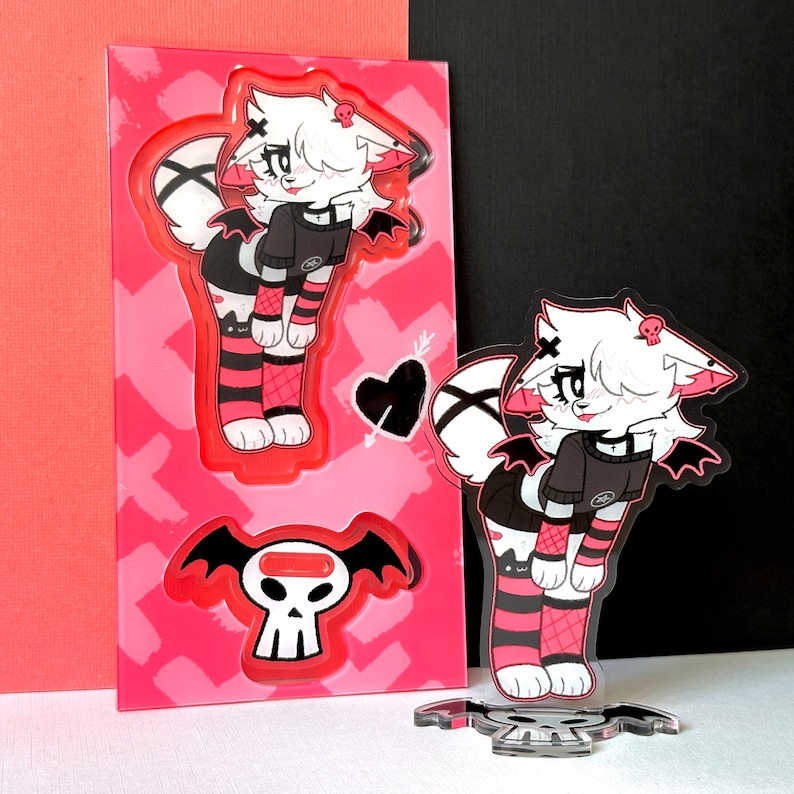 May include: Two acrylic character stands featuring a white and black cat with pink accents wearing a black and white outfit. The cat on the left is standing on a pink and white background with a skull and wings. The cat on the right is standing on a clear base with a skull.