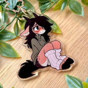 May include: A clear acrylic keychain featuring a cartoon character with black hair, horns, and glasses. The character is wearing a green sweater and white and pink striped socks. A gold-colored clasp is attached to the top.
