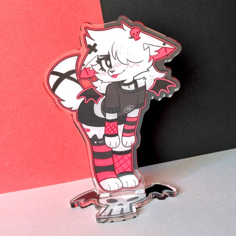 May include: A white and black anime-style character with pink accents and a skull on its head. The character is wearing a black shirt with a white star and black and red striped stockings. The character is standing on a clear acrylic base with a skull design.