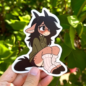 May include: A sticker depicting a cartoon character with goat-like features, wearing a green sweater, glasses, and striped socks. The character has black hair and horns, with a brown tail. The sticker is held against a backdrop of green leaves.