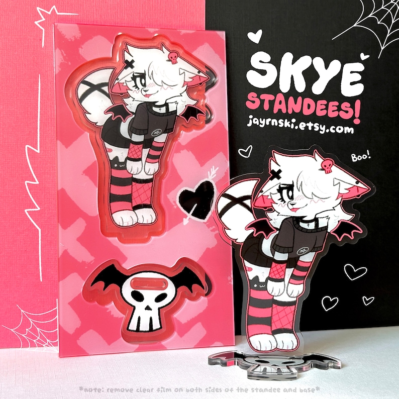 May include: A white furry character with black wings and pink and black striped stockings stands on a skull-shaped base. The character has a black choker and a black shirt with a white X on it. The character is holding a small skull in its right hand. The background is pink with hearts and arrows. The text "SKYE STANDEES! jayrnski.etsy.com Boo!" is visible in the background.