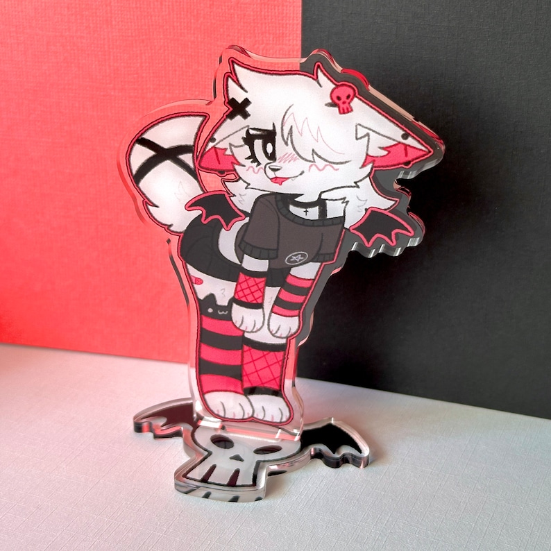 May include: A white and red anime-style character with black wings and red and black striped stockings. The character is wearing a black shirt with a skull and crossbones design. The character is standing on a clear acrylic base with a skull design.