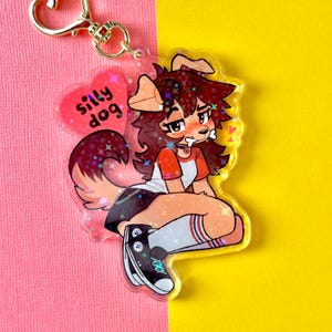 May include: A clear acrylic charm featuring a cartoon dog-like character with brown fur, wearing a red and white shirt, black shorts, and high socks. The charm has a gold-colored clasp and a pink heart with the words "silly dog".