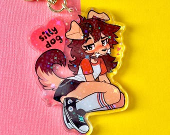 May include: A clear acrylic charm featuring a cartoon dog-like character with brown fur, wearing a red and white shirt, black shorts, and high socks. The charm has a gold-colored clasp and a pink heart with the words "silly dog".