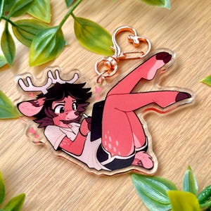 Puede incluir: Un charm de acrílico transparente con un personaje de dibujos animados parecido a un ciervo con cuernos, que lleva una camisa blanca y pantalones cortos negros, sosteniendo una tableta. El charm tiene un cierre en forma de corazón de oro rosa. El personaje es rosa y marrón.