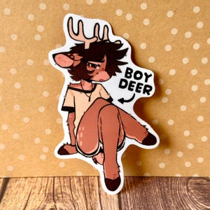 Könnte beinhalten: Ein gestanzter Aufkleber mit einem illustrierten anthropomorphen Hirschcharakter mit braunem Fell, dunklem Haar und Geweih. Die Figur trägt ein helles T-Shirt und sitzt. Der Text BOY DEER ist mit einem darauf zeigenden Pfeil aufgedruckt.