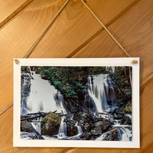 May include: A framed photograph of a waterfall cascading over rocks and lush greenery. The water is flowing rapidly and creates a sense of motion. The image is printed on a white background and framed with a white border.