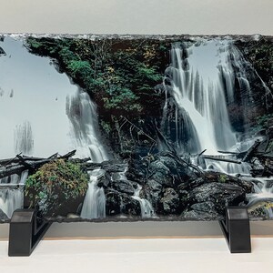 May include: A black slate photo print of a waterfall cascading over rocks and greenery. The print is on a black stand.