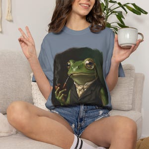 Könnte beinhalten: Hellblaues T-Shirt mit einer detaillierten Illustration eines grünen Frosches in Anzug, Monokel und Pfeife rauchend. Der Frosch ist vor einem dunklen Hintergrund platziert. Das T-Shirt ist ein lässiger, bequemer Stil.