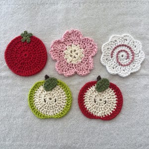 May include: A collection of five handmade crochet coasters. Designs include a red tomato, a pink flower, a white spiral, and two apple slices in green and red. Each coaster is made with a textured stitch.