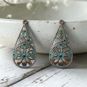 May include: Two teardrop-shaped metal pendants featuring an intricate openwork filigree design with a copper base and a teal patina finish. Each pendant has a small circular loop at the top for attachment, showcasing a floral pattern at the base.