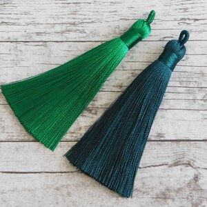 May include: Two green silk tassels, one a bright green and the other a darker teal green. The tassels are on a white wooden background.