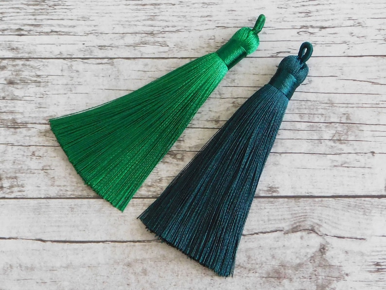 May include: Two green silk tassels, one a bright green and the other a darker teal green. The tassels are on a white wooden background.