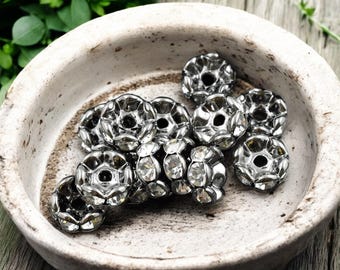 May include: A close-up of a pile of silver-toned metal embellishments with clear rhinestones. The embellishments are in the shape of a flower with a center hole.