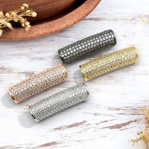 May include: Four cylindrical jewelry beads in silver, gold, rose gold, and gunmetal. Each bead is covered in small, sparkling stones. The beads are arranged on a white wooden surface, with dried flowers and a wooden bowl in the background.