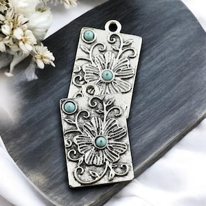 May include: Two silver-toned metal charms with a floral design and turquoise-colored stones. The charms are rectangular and have a loop at the top for hanging.