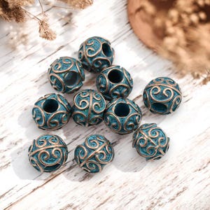 May include: A collection of teal and bronze-colored spherical beads with intricate, swirling patterns. The beads are evenly spaced and appear to be made of metal. The beads are likely for jewelry making or other craft projects.