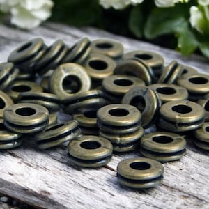 May include: A collection of antique brass-colored metal grommets with a dark, aged finish. The grommets are stacked and scattered, showcasing their circular shape and central hole. These hardware pieces are ideal for various crafting and design projects.