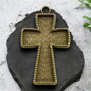 May include: A gold-toned metal cross pendant with a rope-like border. The cross is designed to be filled with resin or other materials.