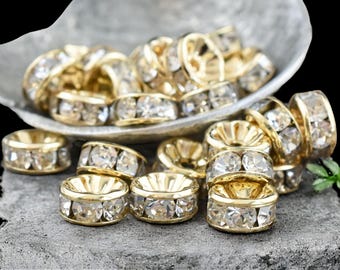 May include: Gold-toned metal beads with clear, faceted crystal accents. The beads are arranged in a decorative display, some in a shallow dish and others on a textured gray surface. The crystals catch the light, creating a sparkling effect.