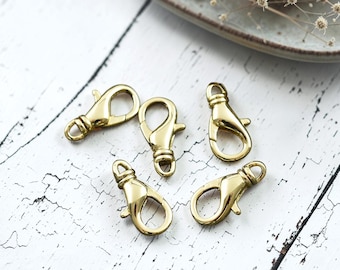 May include: Five gold-toned metal lobster claw clasps arranged in a cluster. Each clasp features a smooth, polished finish, a spring-loaded gate mechanism, and a small circular loop at the base for attachment to jewelry or other craft projects.