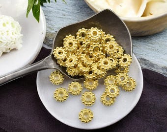 May include: A close-up of a pile of gold-colored metal beads with a raised, circular design. The beads are arranged in a pile on a white plate and a silver spoon.