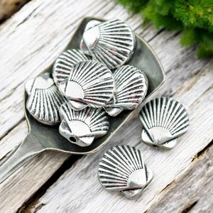 May include: Silver-colored seashell beads with detailed fan-like ridges are displayed in a metal spoon. The beads are scattered on a weathered wooden surface, with a few beads outside the spoon. The image has a rustic, beachy theme.