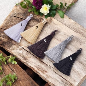 May include: Six different colored tassels with a loop at the top. The tassels are arranged in a row on a wooden surface. The tassels are in the following colors: light purple, beige, dark brown, light gray, black, and black.