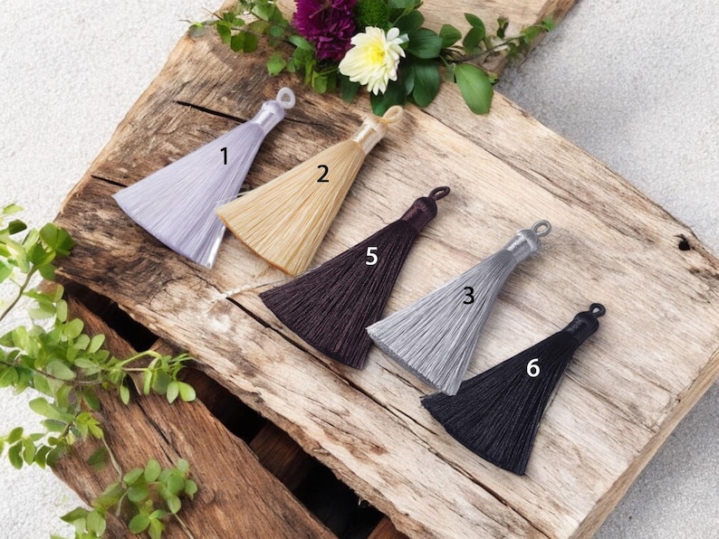 May include: Six different colored tassels with a loop at the top. The tassels are arranged in a row on a wooden surface. The tassels are in the following colors: light purple, beige, dark brown, light gray, black, and black.