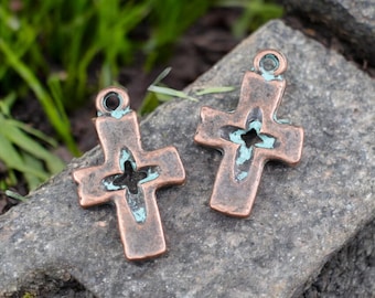 May include: Two bronze-colored cross pendants with a distressed finish and a turquoise patina. Each cross has a cutout design in the center and a small loop at the top for hanging. The crosses are displayed on a gray stone surface.