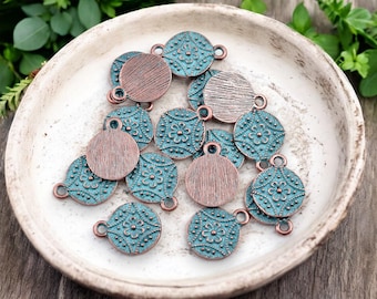 Puede incluir: Una colección de abalorios metálicos redondos y circulares con un acabado turquesa y cobre envejecido. Los abalorios presentan diseños florales y texturizados, con pequeños bucles para sujetarlos a joyas o manualidades. Están dispuestos en un cuenco blanco.