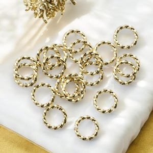 May include: A collection of 15 gold-colored twisted metal rings. The rings are all the same size and are arranged in a random pattern on a white surface.