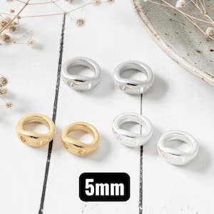 May include: A collection of small, round, ring-shaped beads with two side holes, featuring a smooth metallic finish. The set includes two gold-colored beads and three silver-colored beads, each measuring 5mm in diameter as indicated by the text.