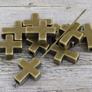 May include: A group of antique gold-toned metal cross-shaped beads with a hole in the centre of each cross. The beads are arranged in a pile on a wooden surface. A thin metal wire is threaded through the beads.