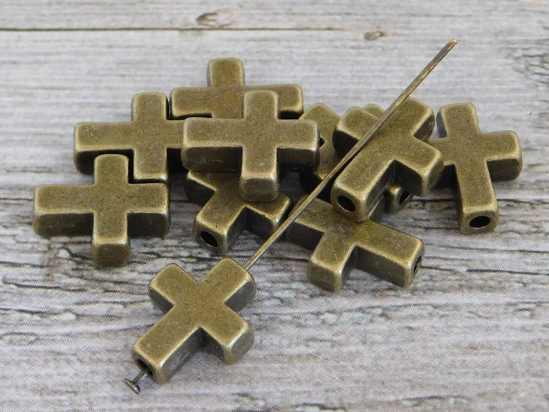 May include: A group of antique gold-toned metal cross-shaped beads with a hole in the centre of each cross. The beads are arranged in a pile on a wooden surface. A thin metal wire is threaded through the beads.