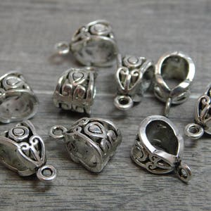 May include: Eight silver-toned metal pendant caps with ornate designs. The caps are designed to be used as a decorative element for necklaces or bracelets.