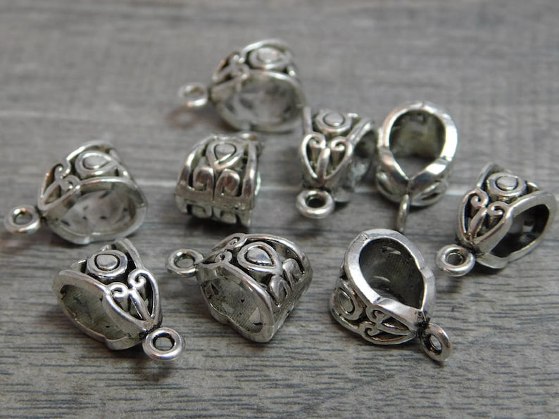 May include: Eight silver-toned metal pendant caps with ornate designs. The caps are designed to be used as a decorative element for necklaces or bracelets.
